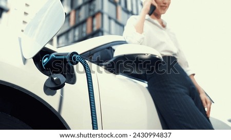 Businesswoman using smartphone, recharge electric car from charging station at residential parking car area. Modern urban lifestyle and eco-friendly electric car utilization to reduce CO2. Peruse