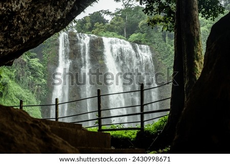 This photo depicts a captivating view of a waterfall cascading down inside a cave. The water appears powerful and refreshing, surrounded by lush green vegetation growing on the cave wallls. Royalty-Free Stock Photo #2430998671