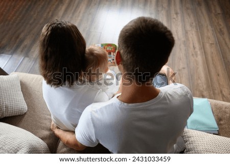 Parents read a book to their child, sitting on the sofa, looking at educational pictures, rear view. Young loving parents spend time with their baby. Concept of happy family, learning.