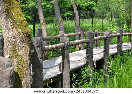 A splitrail fence lines the wooden bridge in the heart of a lush forest, surrounded by water, plants, and grass. The natural landscape is enhanced by the rustic charm of the bridge