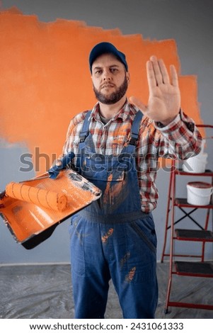 Painter man in blue overalls and cap holding a paint roller on wall background making stop gesture. Copy space