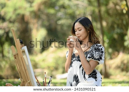 Peaceful young woman sitting in front of easel in the garden and drinking hot coffee