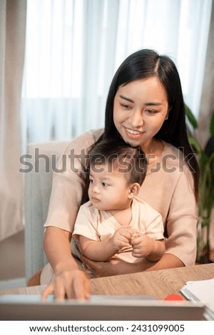 A positive Asian mom is taking care of her baby boy while working on her laptop computer, working remotely from home.