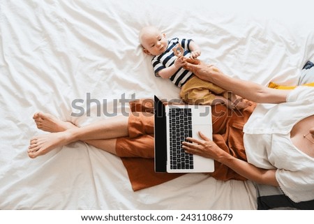 Mother working at laptop playing with baby in bed at home. Modern woman balances between work and baby. Child playing with toy on bed while mother using laptop: surfing internet, doing online shopping