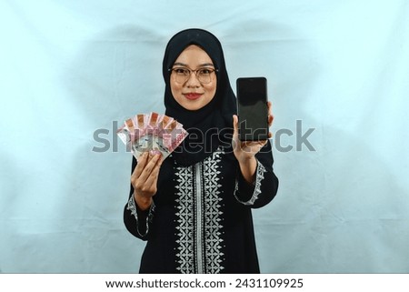 Asian Muslim woman wearing hijab, glasses and black dress with white pattern showing mobile phone with blank screen and holding cash rupiah banknotes isolated on white background