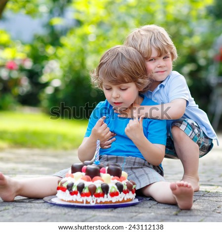 Two little kid boys celebrating birthday in home's garden with big cake.  Two little children having fun with hugging and tasting cake together.