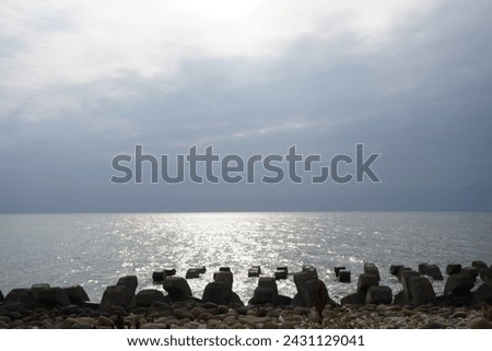 Under the azure sky, fluffy white clouds drift above the coastline, and sunlight sprinkles across the sparkling sea surface. Along the shore, neatly arranged wave breakers create a picturesque scene.  Royalty-Free Stock Photo #2431129041