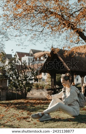 Young Fashionable Teenage Girl With Smartphone In Europian Park In Autumn Sitting At Smiling. Trendy Young Woman In Fall In Park Texting. Retouched, Vibrant Colors. Beautiful Blonde Teenage Girl