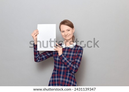 Studio portrait of happy young blond woman holding white blank paper sheet with place for your text and design in hands, smiling cheerfully, wearing checkered dress, standing over gray background