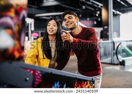 generation z multiracial couple playing pinball machine in the game room Royalty-Free Stock Photo #2431140109