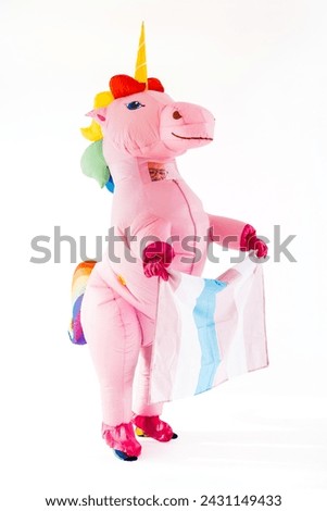 A person disguised in a pink unicorn costume raising a transgender flag in a vibrant and colorful setting. The individual is standing confidently and proudly displaying the flag.