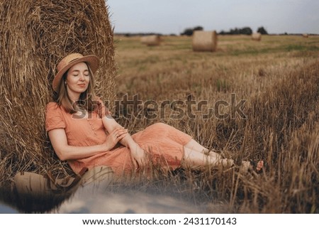 A beautiful girl in a long dress and with long hair a cute smile sitting clouding about a bale of straw. The girl dreamily closed her eyes