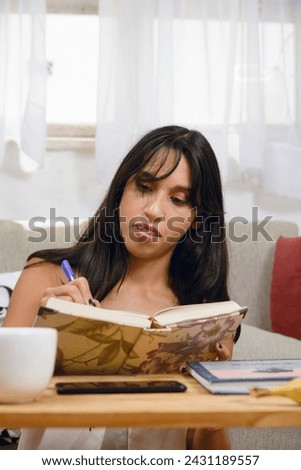 vertical image of young black-haired Argentinian Latina brunette, at home sitting in living room, studying, concentrating, writing down and reading in her notebook