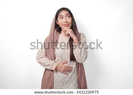 Portrait of Asian Muslim woman wearing headscarf veil placing hands on the stomach feeling hungry and want to eat while fasting, isolated over white background. Ramadan and Eid Mubarak concept Royalty-Free Stock Photo #2431220975