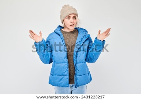 Confused young woman in winter clothes shrugging her shoulders against a white background.