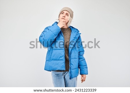 Woman in winter attire with pensive expression, thinks, chooses isolated on light background