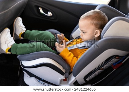 Young boy sitting in baby car booster seat and watching cartoons on mobile phone.