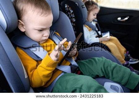 Cute siblings sitting in baby booster car seats and using mobile phones during the road trip. Technology and travel concepts.
