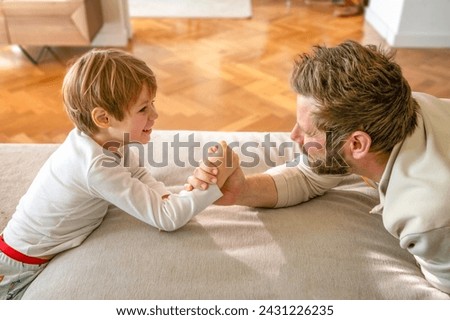 Raising a real man. Concentrated father and son arm wrestling and competing while lying on floor, having fun at home, side view. Family bond, leisure time and fatherhood concept