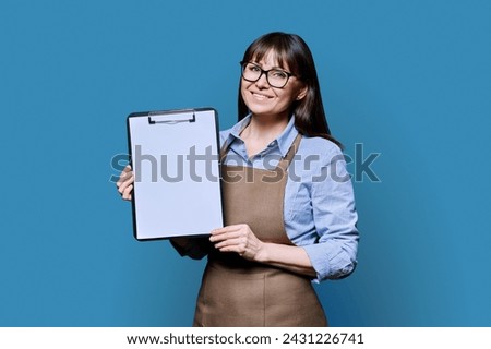 Smiling woman worker, owner in apron showing blank sheet of paper on clipboard Royalty-Free Stock Photo #2431226741
