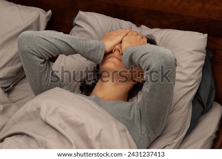 A young woman lying in bed covering her face with her hands; an ill or sick female is finding it hard to cope; a stressed or anxious lady lying in bed doesn’t want to get up
 Royalty-Free Stock Photo #2431237413