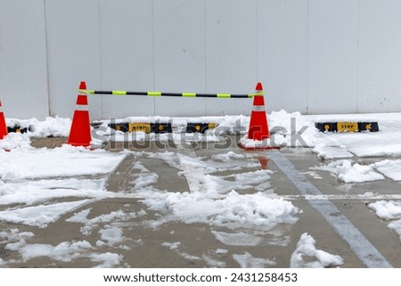 Bright orange traffic cones standing on car park, security barrier with snow. Plastic reflective road cones on snowy winter urban street against cars parking