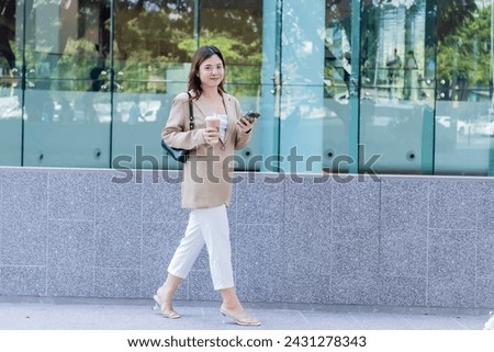 Young asian elegant business woman leader wearing suit walking near modern city building using cell phone platform applications and hold coffee drink walking on street outdoors.