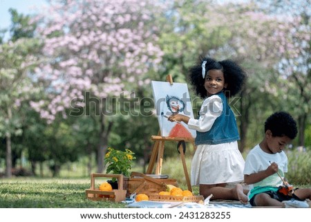 African little girl holds brush and paints picture on easel at park, very happy and smiling at camera, her younger brother sat next to her