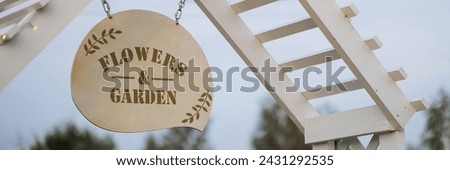 A round Flowers and Garden sign hangs from a white, wooden structure. Concept for garden beautification and outdoor events. Banner with copy space.