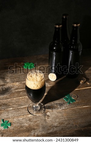 Bottles and glass of dark beer, coins and clovers for St. Patrick's Day celebration on wooden table against black background