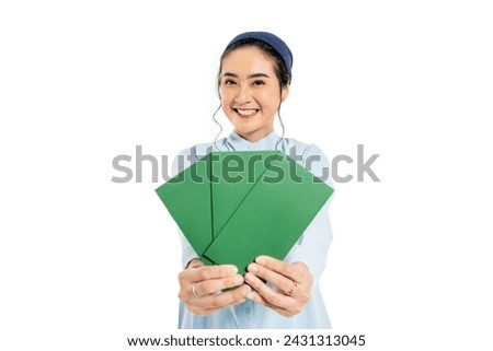 Smiling young beautiful Asian muslim wearing hijab showing envelope of money for eid al fitr present THR looking at camera. wearing modern ramadan outfit for eid mubarak idul fitri Royalty-Free Stock Photo #2431313045