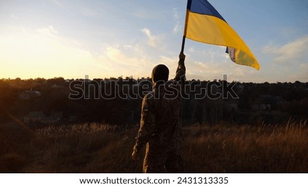 Male soldier stands with raised flag of Ukraine against background of beautiful sunset. Male ukrainian army soldier with a lifted blue-yellow banner in honor of the victory against russian aggression