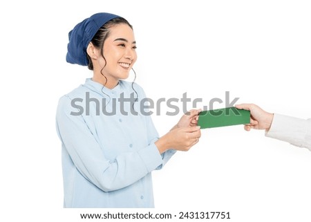 Smiling young beautiful Asian muslim wearing hijab receiving envelope of money for eid al fitr present THR. wearing modern ramadan outfit for eid mubarak idul fitri Royalty-Free Stock Photo #2431317751