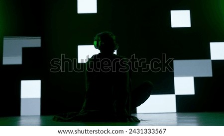 Person against big digital wall in studio. Man in virtual reality glasses sitting in front of digital screen cyber graphic visual background, back shot.