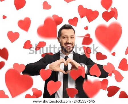 Young man in a suit and tie gesturing a heart symbol and standing under falling hearts isolated on white background