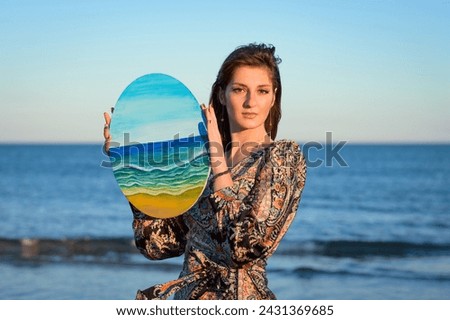 A woman wearing a dress holds a painted picture in her hand, showcasing the colorful design. She gazes at the picture, displaying a sense of appreciation for the artwork. Royalty-Free Stock Photo #2431369685