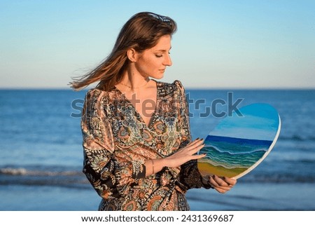 A woman wearing a dress holds a painted picture in her hand, showcasing the colorful design. She gazes at the picture, displaying a sense of appreciation for the artwork.