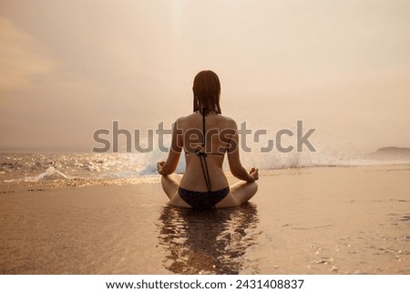 A woman in a swimsuit adopts the lotus position, practicing yoga on the beach at sunset Royalty-Free Stock Photo #2431408837