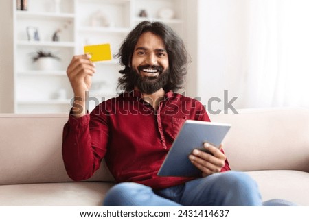 Positive young indian man with digital tablet and credit card sitting on couch at home, paying for goods and services online, copy space. Easy banking, Internet shopping, e-commerce Royalty-Free Stock Photo #2431414637