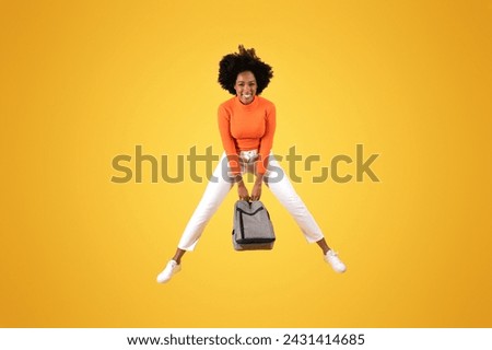 Dynamic overhead shot of a delighted young african american woman with curly hair, holding a backpack between her legs, dressed in an orange shirt and white pants on a yellow background