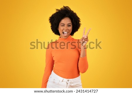 Vivacious young african american woman with fluffy hair winks and flashes a peace sign, clad in a snug orange turtleneck and crisp white jeans, against a monochromatic yellow background Royalty-Free Stock Photo #2431414727