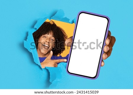 Joyful young woman with curly hair laughs as she points to a blank smartphone screen while breaking through a blue torn paper background. Recommendation website, app, good news, win and technology