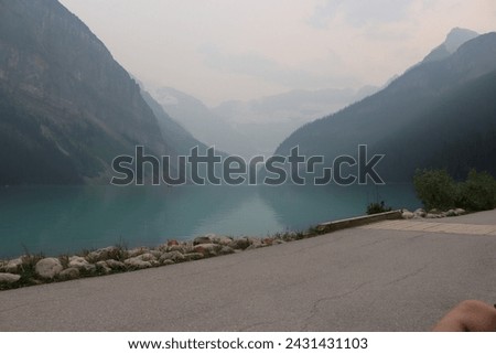 The image depicts a road running alongside a body of water with mountains in the background, taken at Lake Louise. The scenery includes a fjord, fog, and a mountain range, creating a pictures