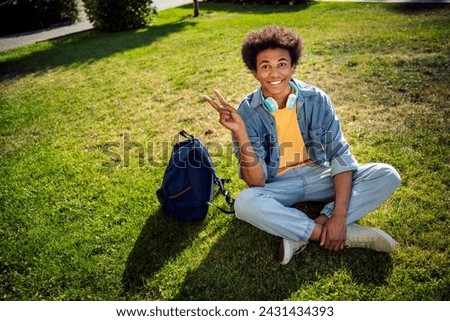 Photo of good mood cheerful guy wear denim jacket earphones relaxing lawn showing v-sign outside urban city street