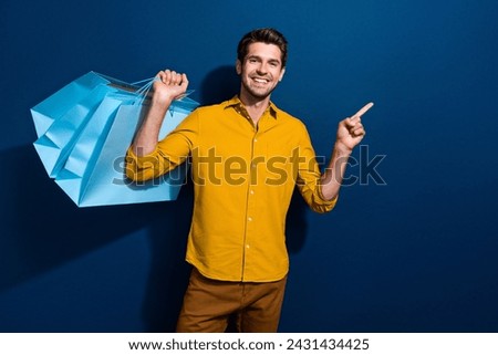 Photo portrait of attractive young man shopper bags point empty space dressed stylish yellow clothes isolated on dark blue color background