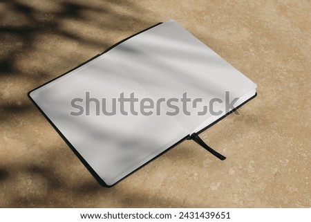Tropical summer mockup. Open diary, book with black cover in sunlight. Blank pages. Harsh tree branches shadows. Beige marble stone texture, background. Holiday template. Flat lay. Top view, no people