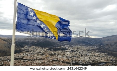 State flag of Bosnia and Herzegovina waving on the wind. Bosnia Herzegovina national flag blowing on flagpole above the city of Mostar, aerial drone view.  Royalty-Free Stock Photo #2431442349
