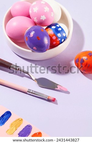 Closeup view of a white dish containing beautiful Easter eggs, displayed with a palette knife, a paintbrush and a paper of variety paint colors. Promotion and shopping template for Easter