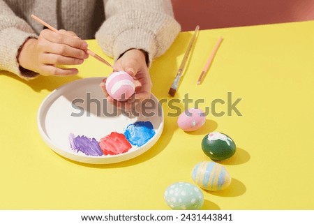 A woman wearing a sweater is painting an Easter egg by a paintbrush, on the dish featured three colors of paint. Some Easter eggs with different patterns are displayed for Easter concept