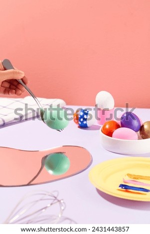 A hand is holding a fork with an easter egg in pastel green color create a reflection in the mirror below. A dish of colorful Easter eggs displayed with a dish of paint colors on the table